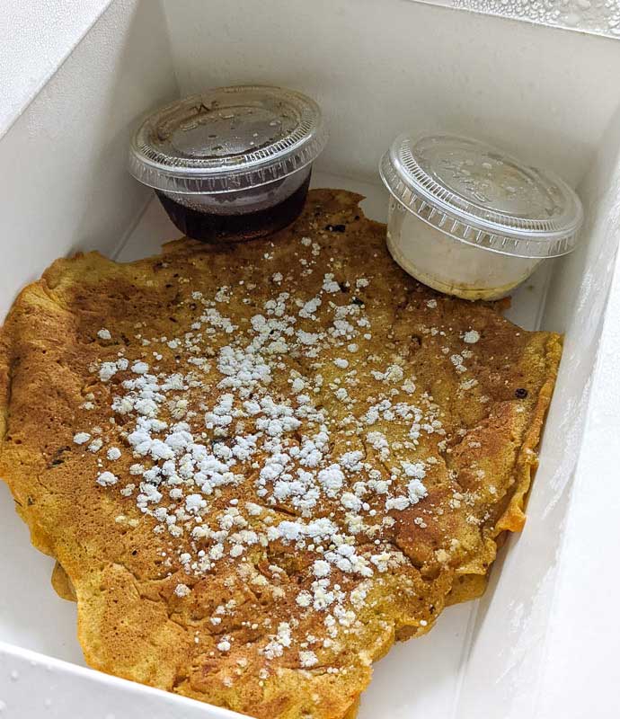 Cardamom pancake with a light dusting of powdered sugar in a takeout box with a side of syrup and cardamom cream cheese